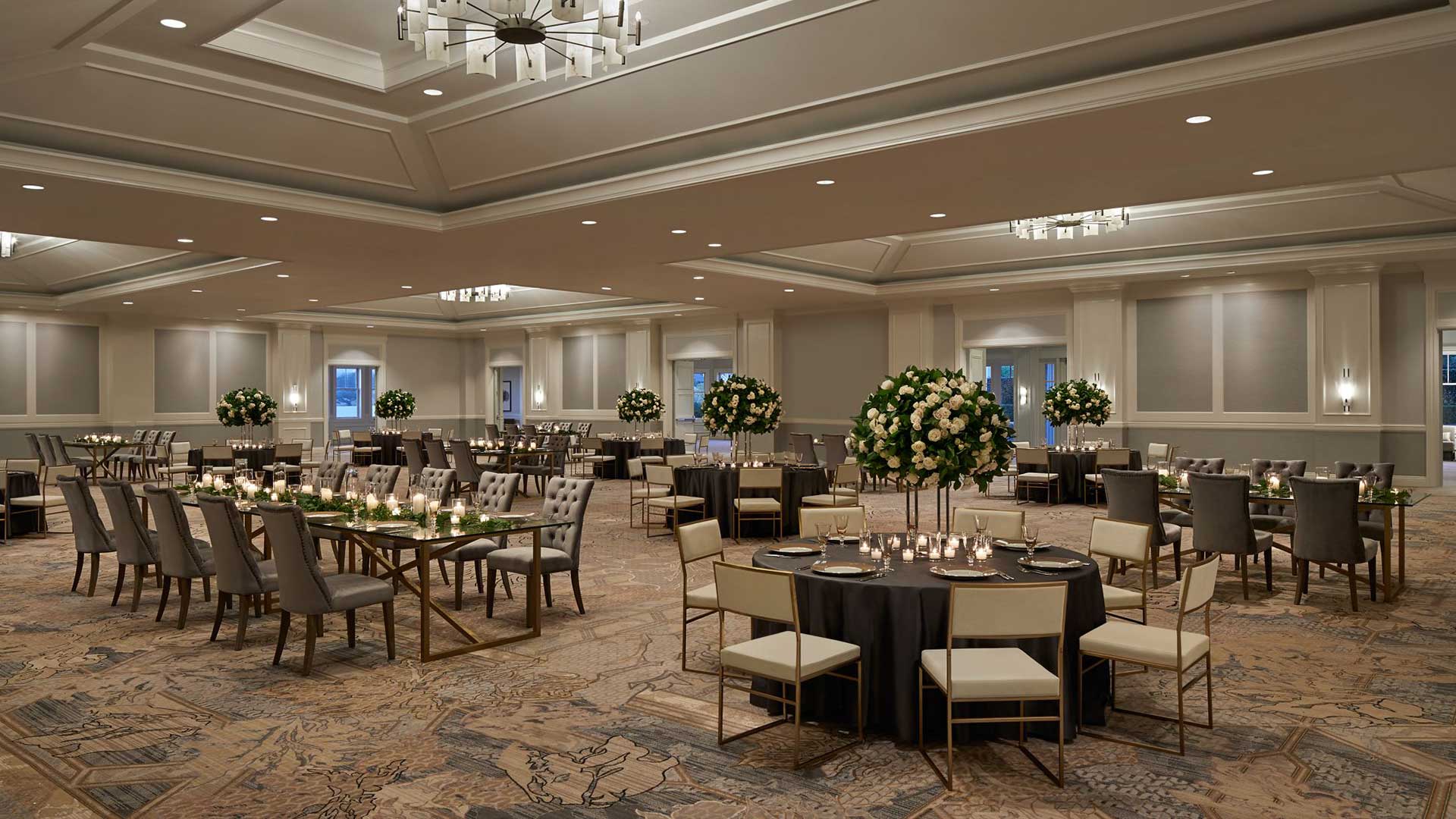 A beautifully decorated banquet hall set up for an event, featuring round tables with black tablecloths, elegant floral centerpieces, and place settings with candles. The room has a neutral color scheme with grey walls, high ceilings, and modern chandeliers. The seating arrangement includes cushioned chairs around the tables and long dining tables with upholstered chairs.