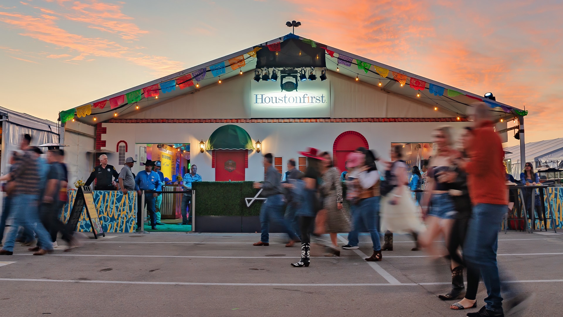 A bustling scene at HoustonFirst's rodeo tent, with people walking by and engaging in activities. The tent is decorated with colorful banners and lights, creating a festive atmosphere. The sign above the entrance reads 'HoustonFirst,' and the exterior features vibrant colors and welcoming decor. The sky is painted with warm hues from the setting sun, adding to the lively ambiance of the event.