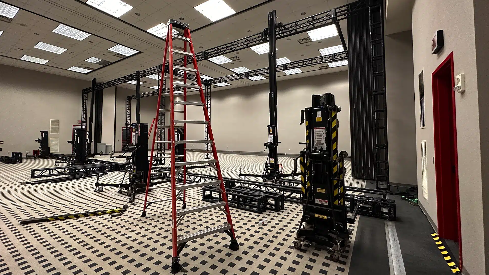 A setup in progress for an event production with various rigging equipment, a tall red ladder, and black truss structures in a spacious room with checkered flooring. The room is well-lit with ceiling lights and has black and yellow safety tape marking areas on the floor.
